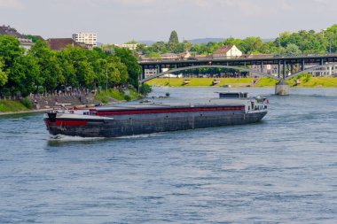 Basel şehri. Rhein nehri boyunca uzanan bir köprü ve arka planda bir şehir var. Köprü üzerinde kırmızı ve beyaz işareti olan taştan bir yapıdır. Şehir binalar ve bir kiliseyle dolu. Gökyüzü açık ve mavi