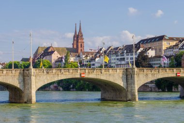 Basel şehri. Rhein nehri boyunca uzanan bir köprü ve arka planda bir şehir var. Köprü üzerinde kırmızı ve beyaz işareti olan taştan bir yapıdır. Şehir binalar ve bir kiliseyle dolu. Gökyüzü açık ve mavi
