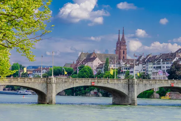 Basel şehri. Rhein nehri boyunca uzanan bir köprü ve arka planda bir şehir var. Köprü üzerinde kırmızı ve beyaz işareti olan taştan bir yapıdır. Şehir binalar ve bir kiliseyle dolu. Gökyüzü açık ve mavi