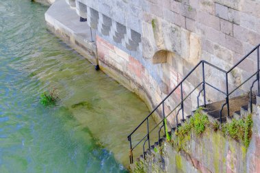 Basel şehri. Rhein nehri boyunca uzanan bir köprü ve arka planda bir şehir var. Köprü üzerinde kırmızı ve beyaz işareti olan taştan bir yapıdır. Şehir binalar ve bir kiliseyle dolu. Gökyüzü açık ve mavi