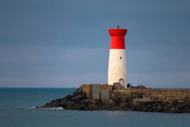 Bir deniz feneri kayalık bir kıyı şeridinde, okyanus dalgaları kayalara çarpıyor. Deniz feneri kırmızı ve beyazdır ve kayalık bir kıyı şeridiyle çevrilidir. Sahne sakin ve huzurlu.