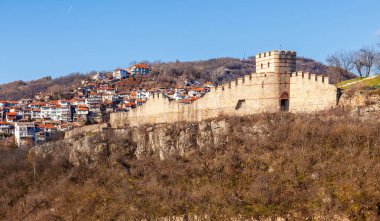 Arka planda bulutlu bir gökyüzü olan tepedeki Çarevets Kalesi. Kale ağaçlar ve kayalarla çevrili.