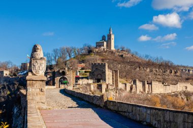 Arka planda bulutlu bir gökyüzü olan tepedeki Çarevets Kalesi. Kale ağaçlar ve kayalarla çevrili.