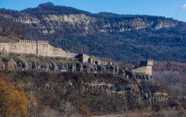 Arka planda bulutlu bir gökyüzü olan tepedeki Çarevets Kalesi. Kale ağaçlar ve kayalarla çevrili.
