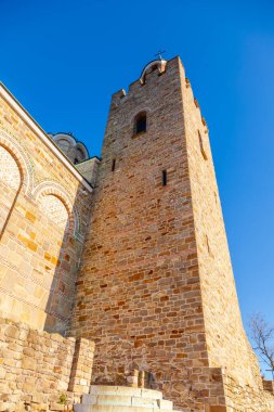 Çan kulesi ve üzerinde haç olan büyük taştan bir bina. Bina ağaçlarla çevrili ve sakin bir atmosferi var.