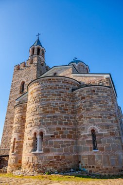 Çan kulesi ve üzerinde haç olan büyük taştan bir bina. Bina ağaçlarla çevrili ve sakin bir atmosferi var.