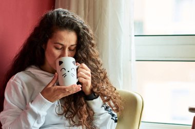 Woman relaxing in a armchair drinking a cup of tea indoors surrounded by plants
