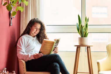 Woman relaxing in a armchair reading a book indoors surrounded by plants