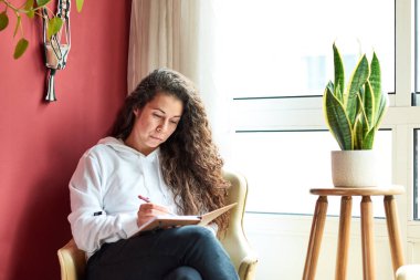 Woman relaxing in a armchair writing a diary indoors surrounded by plants