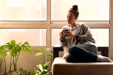 Woman drinking a cup of tea in a couch indoors surrounded by plants