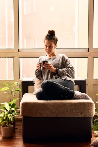 Woman drinking a cup of tea in a couch indoors surrounded by plants