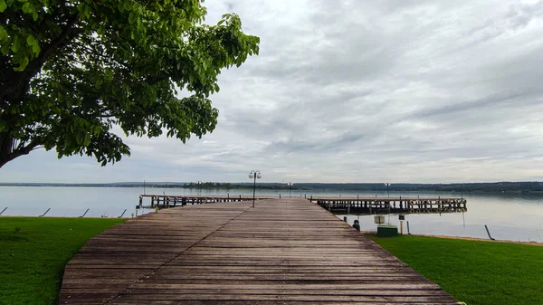 stock image A peaceful atmosphere under the cloudy sky around the lake with wooden dock, meadow and tree viewed