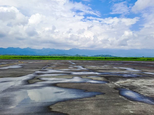 View of concrete floor with puddles after rain, green meadow, mountains and cloudy sky background. Selective focus image