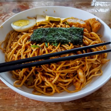 Stir fried noodles with chicken egg, tofu and seaweed topping, served on white bowl with chopsticks. Asian style food. Close up, selective focus