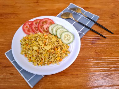 Domates, salatalık ve krakerle birlikte beyaz bir tabakta servis edilen körili pilav. Arka planda doğal ahşap.