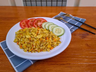 Domates, salatalık ve krakerle birlikte beyaz bir tabakta servis edilen körili pilav. Arka planda doğal ahşap.