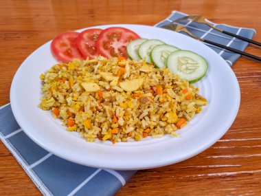 Domates, salatalık ve krakerle birlikte beyaz bir tabakta servis edilen körili pilav. Arka planda doğal ahşap.