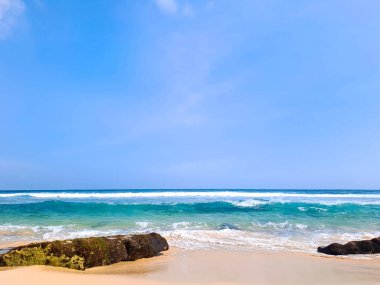 Açık mavi gökyüzü altında beyaz kum, kaya ve sakin dalgalarla güzel bir sahil manzarası. Yaz tatili kavramı. Ngudel Plajı, Malang, Endonezya