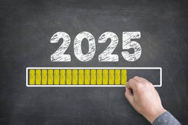 Tebeşir tahtası 2025 'i gösteriyor. Yükleme çubuğuyla mutlu yıllar. Bir el yazarken görülüyor, bayram vesilesiyle yeni yıla geri sayım başlatılıyor..