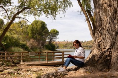 Gölün kıyısındaki bir ağacın altında kitap okuyan kız.