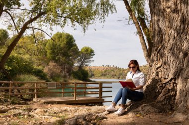 Gölün kıyısındaki bir ağacın altında kitap okuyan kız.