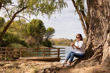 Kız gölün kıyısındaki bir ağacın altında telefonla konuşuyor.