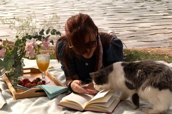 Kedi ve piknik dekorasyonu, çiçekler, meyve ve meyve suyu ile gölün kıyısında kitap okuyan bir kız.