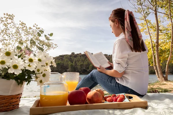 Kız, gölün kıyısında çiçeklerle ve meyvelerle piknik hakkında kitap okuyor.