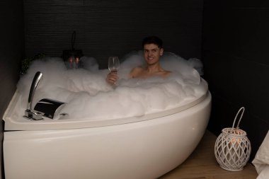 Kaplıcadaki genç adam köpük banyosu yapıyor ve bir bardak şarap içiyor.