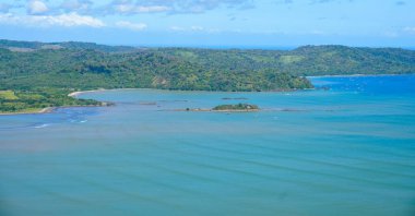 Berrak bir gökyüzünün altında yemyeşil tepeler ve canlı mavi bir okyanus olan güzel bir kıyı şeridi manzarası. Tropikal cenneti ve sakin doğal manzaraları tasvir etmek için mükemmel.