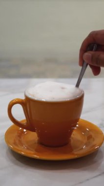 Portakallı kahve fincanında köpüklü bir cappuccino karıştıran elin dikey yavaş hareketi