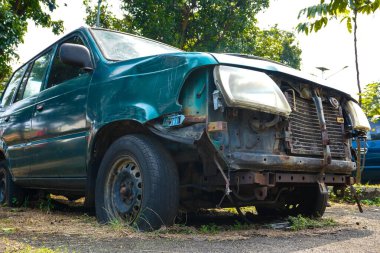 İhmal edilmiş yeşil bir araba aşırı büyümüş bir arsada oturur ve güneş altında lastikleri patlak, ön parçaları eksik izler gösterir. Çevreleyen bitki örtüsü, olay yerinin çürümesine neden olur.