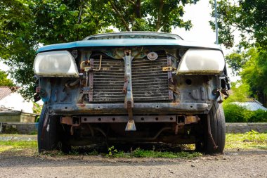 İhmal edilmiş yeşil bir araba aşırı büyümüş bir arsada oturur ve güneş altında lastikleri patlak, ön parçaları eksik izler gösterir. Çevreleyen bitki örtüsü, olay yerinin çürümesine neden olur..
