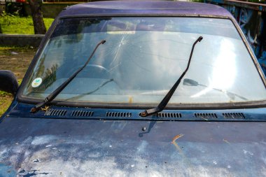 Terk edilmiş bir araçta kırık bir ön cam ve paslanmış bir vücutta aşınma izleri var. İhmal edilmiş otopark uzun otlar ve dağınık molozlarla dolu.