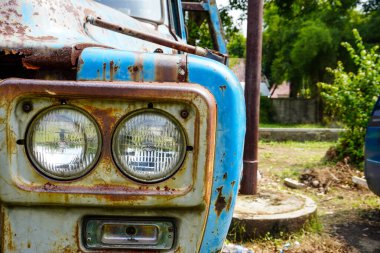 Pas lekeleri ve sisli farları olan yıpranmış mavi bir kamyon ağaçlar ve açık hava elementleriyle çevrili çimenli bir arazide duruyor.