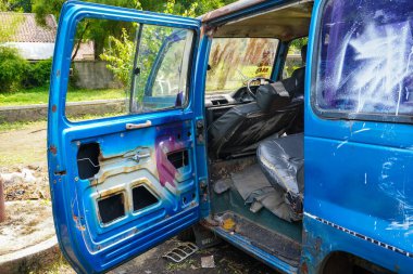 Açık kapısı olan mavi bir aracın yakın çekimi. Pas sergiliyor, boya soyuyor ve dışarıdaki yeşilliklerle çevrili bir ortamda giyiliyor.
