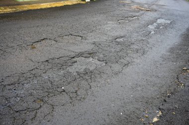 Çukur ve çatlaklardan görünür hasar gören asfalt bir yol, zamanla aşınmayı ve yırtılmayı yansıtıyor. Görüntü hasarlı yüzeyin engebeli dokularını yakalar, bozulma ve zarar görmüş koşulları vurgular.