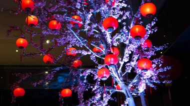 Şenlikli kırmızı fenerler çiçek açan dekoratif bir ağacı süslüyor, bayram veya olay dekorasyonu için mükemmel bir kutlama atmosferi yaratıyor..