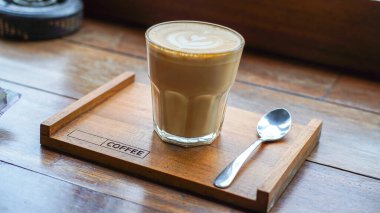 Latte sanatıyla bir bardak kahve, ahşap bir tepside, bir kaşıkla, ahşap bir masada oturur; kafe ve içkiler için mükemmeldir..