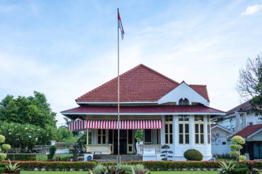 Bengkulu, 20 Eylül 2024: Endonezya bayrağının bulutlu bir gökyüzünde dalgalandığı beyaz bir koloni binası mimarlık ve seyahat projeleri için mükemmel bir manzara oluşturuyor..