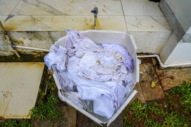 Kirli beyaz kıyafetlerle dolu plastik bir çamaşır sepeti dışarıda, su musluğu ve çimenlerin yanında temizlik ve angarya projelerinde kullanılır..