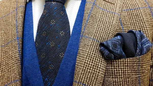 Elegant men's smart casual fashionable outfit, showcasing a brown checkered suit, a dark blue tie with floral pattern and a matching pocket square, accompanied by a blue sweater vest