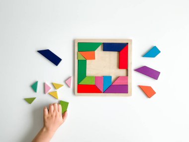 Childs el, beyaz arka planda çok renkli ahşap mozaik koleksiyonu yapıyor. Çocuk renkli bir tangram çözüyor. Beyaz arkaplanda renkli geometrik şekillerin karesi.