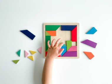 Childs el, beyaz arka planda çok renkli ahşap mozaik koleksiyonu yapıyor. Çocuk renkli bir tangram çözüyor. Beyaz arkaplanda renkli geometrik şekillerin karesi.