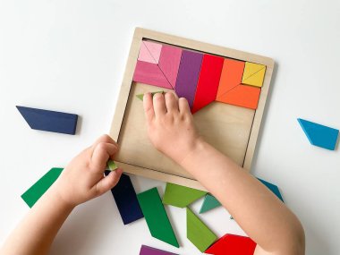 Childs el, beyaz arka planda çok renkli ahşap mozaik koleksiyonu yapıyor. Çocuk renkli bir tangram çözüyor. Beyaz arkaplanda renkli geometrik şekillerin karesi.