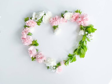 Beyaz arka planda pembe ve beyaz karanfillerden, yeşil yapraklardan, çingenelerden oluşan bir kalp şeklinde çerçevelenmiş. Düz yerde, üst tarafta. Bahar arka planı. Düğün, kartlar ve davetiyeler için uygun