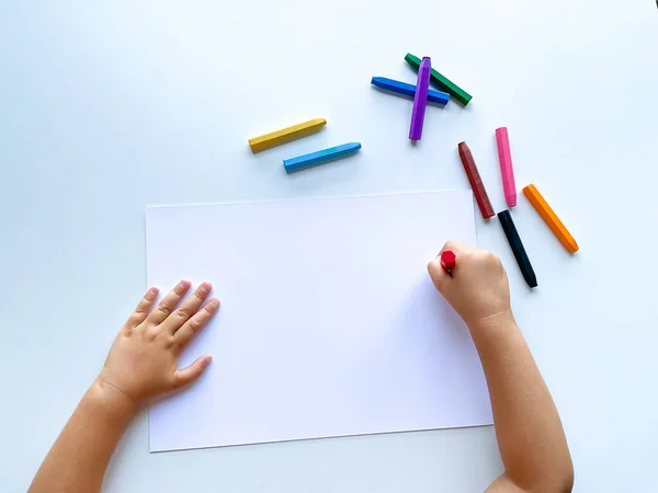 Çocukların elleri beyaz bir kağıt yaprağına renkli mum boya kalemleriyle çizer. Boş bir sayfanın üst görüntüsü. Yüksek kalite