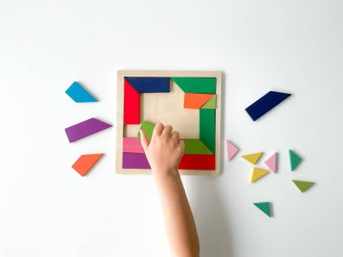 Childs el, beyaz arka planda çok renkli ahşap mozaik koleksiyonu yapıyor. Çocuk renkli bir tangram çözüyor. Beyaz arkaplanda renkli geometrik şekillerin karesi.