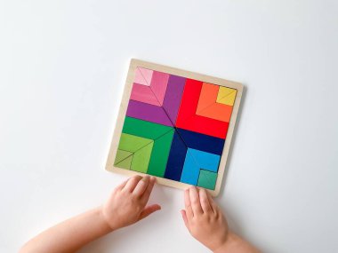 Childs el, beyaz arka planda çok renkli ahşap mozaik koleksiyonu yapıyor. Çocuk renkli bir tangram çözüyor. Beyaz arkaplanda renkli geometrik şekillerin karesi.