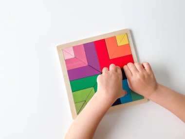 Childs el, beyaz arka planda çok renkli ahşap mozaik koleksiyonu yapıyor. Çocuk renkli bir tangram çözüyor. Beyaz arkaplanda renkli geometrik şekillerin karesi.